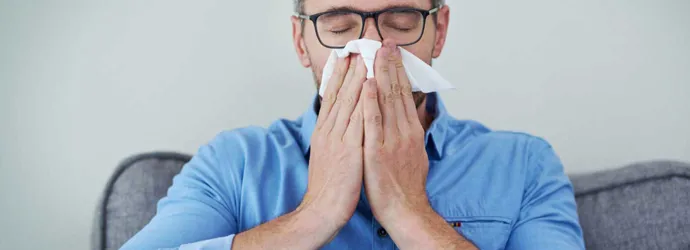 Un hombre con gafas y una camisa azul sentado en un sofá sonándose la nariz