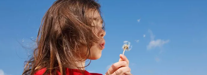 Uma criança pequena que pode precisar de alguns remédios caseiros para a febre dos fenos enquanto sopra um dente-de-leão com um céu azul de fundo