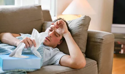 Homem deitado no sofá com gripe enquanto segura lenços de papel