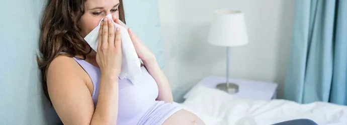 Uma mulher grávida sentada numa cama a assoar o nariz com um lenço de papel