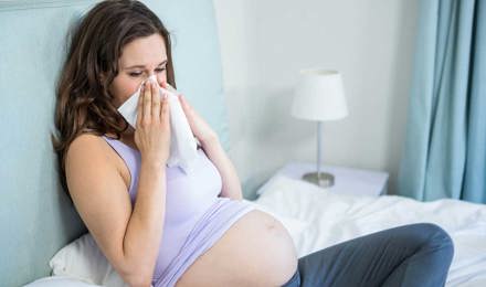 Uma mulher grávida sentada numa cama a assoar o nariz com um lenço de papel